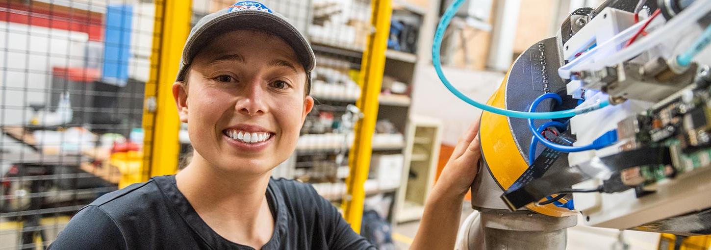 在机器人实验室里，戴着NASA球帽的伊利诺伊大学学生微笑着把手放在一个大型机器人的手臂上.